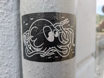 Street sticker The sticker depicts an abstract, stylized octopus or kraken-like sea creature with swirling tentacles, rendered in light-colored (possibly white or silver) lines against a solid black background. Several small, simple fish shapes are scattered around the main creature. The sticker is placed on a textured, light-colored pole or column, showing some signs of wear and scratches on its surface.