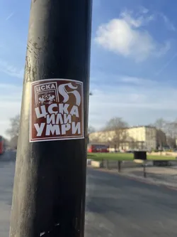 Street sticker A rectangular sticker with a dark reddish-brown background and a thin white border, affixed to a black pole. The primary text and imagery are rendered in white. At the top left, within a smaller white-bordered rectangle, is the Cyrillic acronym 'ЦСКА' above the words 'ФУТБОЛЕН КЛУБ' (Football Club). Below this, a stylized skull with a football (soccer ball) in its mouth is depicted. To the right of the top section, a large, stylized white 'S' or similar ornate character is present. The dominant text at the bottom, in large, bold Cyrillic letters, reads 'ЦСКА ИЛИ УМРИ' (CSKA or Die).