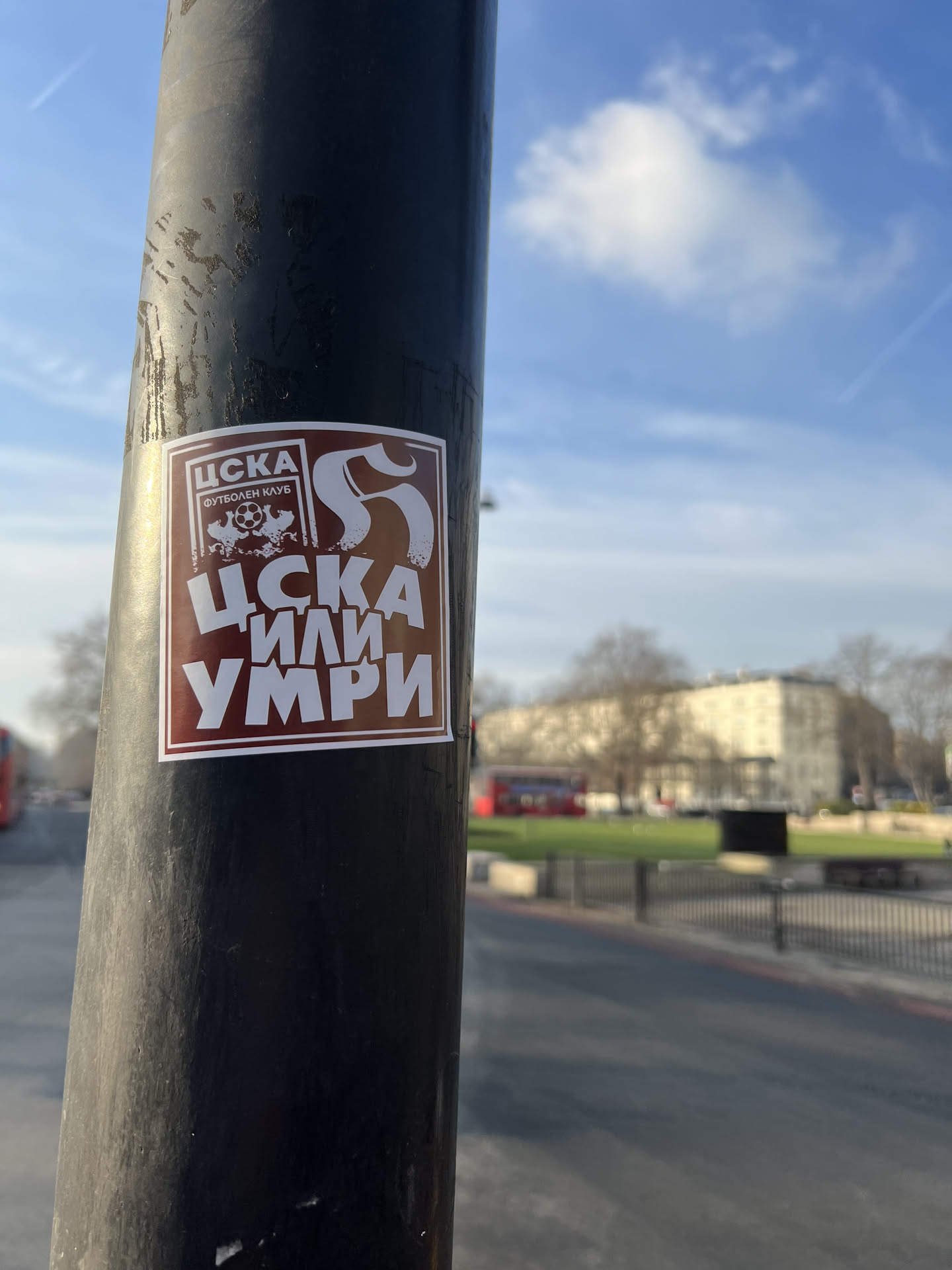 A rectangular sticker with a dark reddish-brown background and a thin white border, affixed to a black pole. The primary text and imagery are rendered in white. At the top left, within a smaller white-bordered rectangle, is the Cyrillic acronym 'ЦСКА' above the words 'ФУТБОЛЕН КЛУБ' (Football Club). Below this, a stylized skull with a football (soccer ball) in its mouth is depicted. To the right of the top section, a large, stylized white 'S' or similar ornate character is present. The dominant text at the bottom, in large, bold Cyrillic letters, reads 'ЦСКА ИЛИ УМРИ' (CSKA or Die).