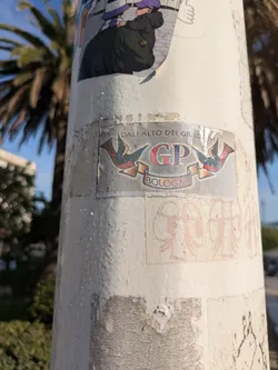 Street sticker The sticker is affixed to a white pole. The sticker features a design with two swallows facing each other, with their wings forming an arch. Between the swallows, the letters 'GP' are prominently displayed in a bold, red and gray font. Below the letters, the word 'BOLOGNA' is written on a banner. Above the swallows is the text 'DALL'ALTO DEI GRADINI'. The sticker appears aged and worn.