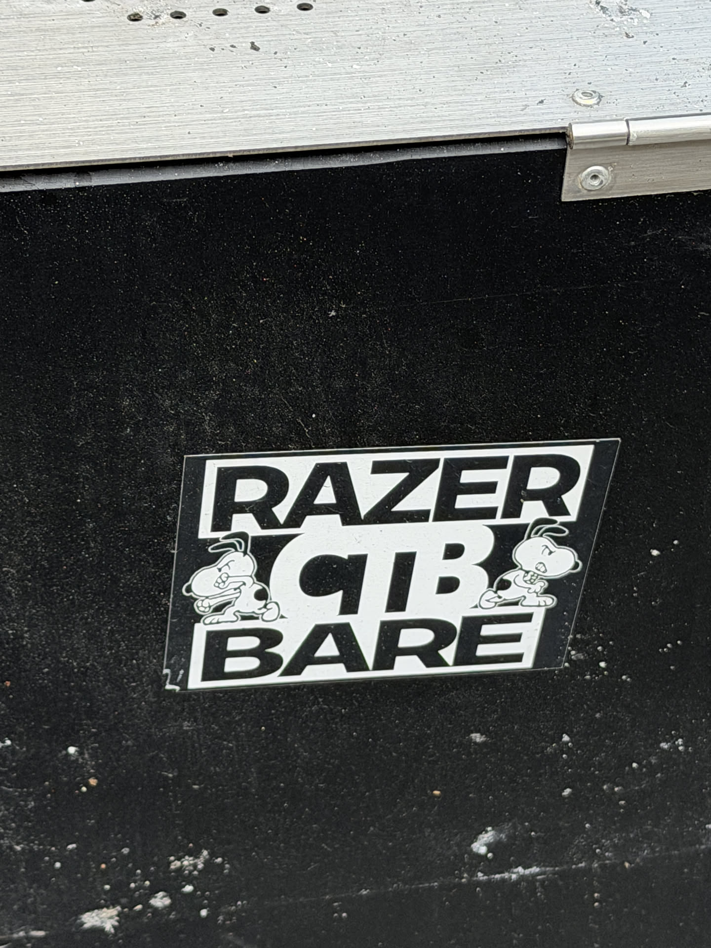 A rectangular sticker with a black background and white design elements, stuck onto a textured black surface. The sticker displays bold white text in three rows: 'RAZER' at the top, 'CTB' in the middle, and 'BARE' at the bottom. On either side of the central 'CTB' text are mirrored illustrations of an angry, muscular cartoon dog resembling Snoopy, shown with clenched fists and a fierce expression.