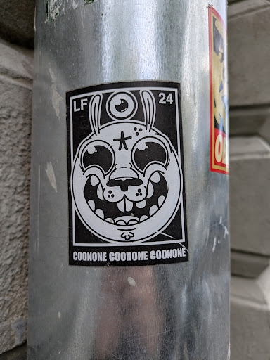 Street sticker This sticker has a black background and white ink. The design is a cartoon-style rabbit head with exaggerated features. It has a third eye on its forehead, a star on its face, and a wide, toothy grin. The text "COONONE COONONE COONONE" is printed at the bottom of the sticker. It is affixed to a silver pole.