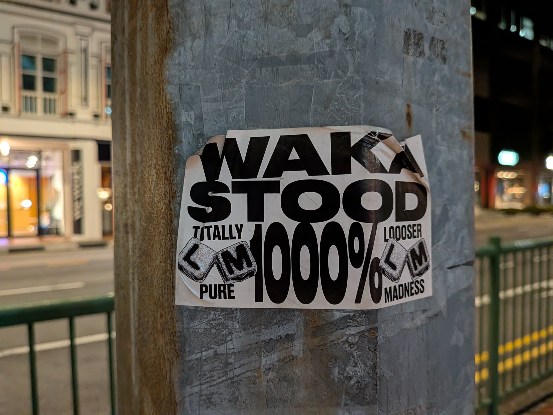 A rectangular black and white sticker on a metal pole. It features bold, stylized text reading 'WAKA STOOD' at the top and '1000%' in the center. The sides contain the phrases 'TOTALLY PURE' and 'LOOOSER MADNESS' accompanied by small illustrations of cubes labeled 'L' and 'M'.