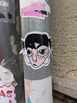 The sticker features a cartoon girl with black hair in pigtails, green eyes, and a fair complexion. Her expression is apathetic. The sticker is placed on a gray pole with other stickers.