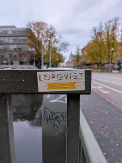 Street sticker The sticker is rectangular with a light-colored background, appearing to be made of paper. It is affixed to a dark green metal railing. The top portion of the sticker features the word 'LOFQVIST' in a clean, sans-serif font with black lettering. The 'O' in 'LOFQVIST' is stylized to resemble a circle with a line through it, and the 'Q' has a similar circular element. Below the text, there is a horizontal band of bright yellow, resembling tape, with a pointed end on the right side. The edges of the sticker show signs of wear and slight peeling, indicating it has been placed outdoors.