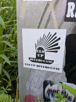 Street sticker The sticker is white with a central black graphic resembling a sun or a rising symbol with rays. Inside the base of this symbol, the words "GLO UP" are printed vertically. Below the symbol, the words "DINERO GANG" are written horizontally. The full phrase "GLO UP DINERO GANG" is repeated again in black text at the bottom edge of the sticker. The sticker is placed on a light grey pole, with green foliage visible around it.