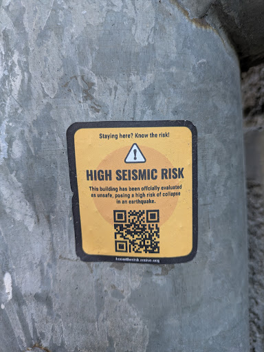 A square sticker with rounded corners on a pole. The sticker is yellow with a black border. The text on the sticker indicates that the building has been officially evaluated as unsafe, posing a high risk of collapse in an earthquake. There is a QR code at the bottom of the sticker and the text "Staying here? Know the risk!"