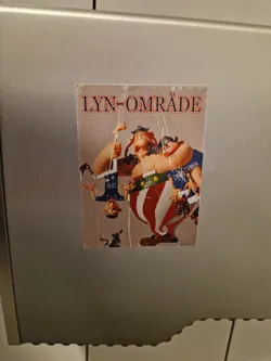 Street sticker A sticker depicting a humorous cartoon scene of vikings. The text "LYN-OMRÅDE" is visible at the top.  The style is reminiscent of claymation or stop-motion animation. The sticker appears to be affixed to a metallic surface, possibly in a public restroom.