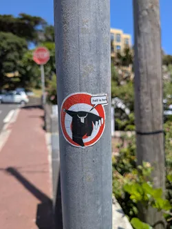 Street sticker A weathered circular sticker on a metal pole. It depicts a black silhouette of a priest running against a red and white circular background, with a speech bubble reading 'hell is here'.