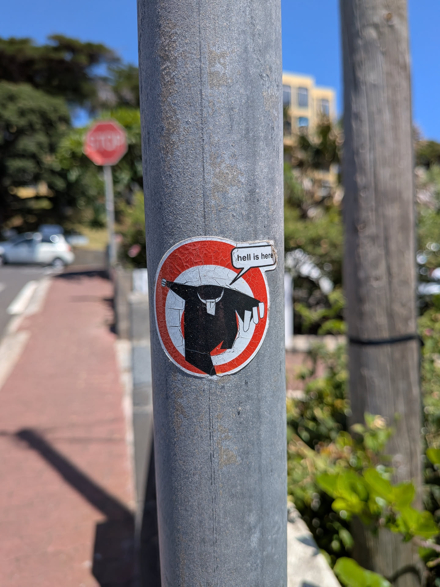 A weathered circular sticker on a metal pole. It depicts a black silhouette of a priest running against a red and white circular background, with a speech bubble reading 'hell is here'.