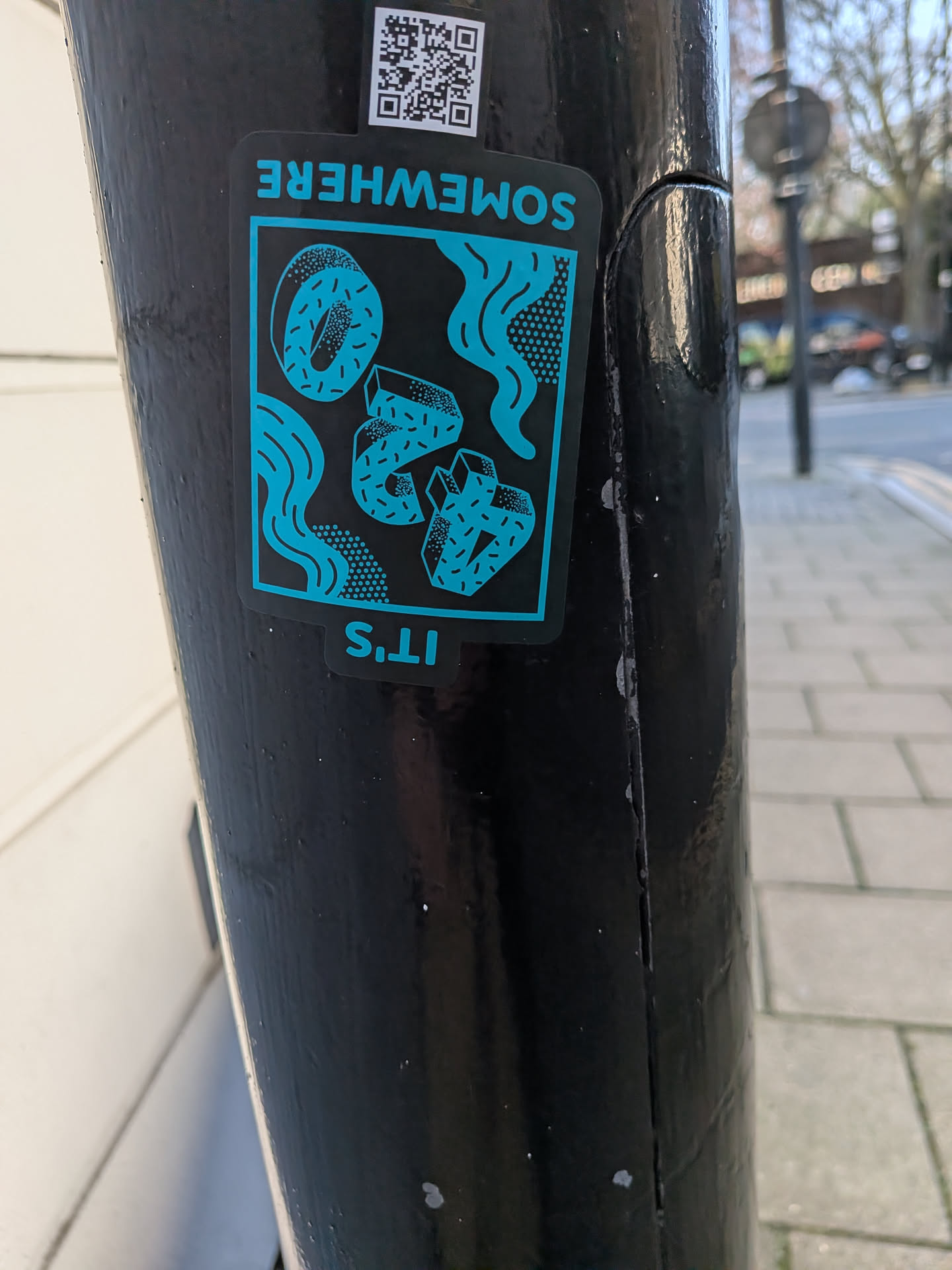 A rectangular sticker with rounded corners, featuring a striking two-tone design in black and a bright teal/cyan. The background is black, while all illustrative elements are rendered in teal. The central image depicts a surreal composition of objects: a whole donut with sprinkles in the upper left, two stylized or broken donut-like shapes with sprinkles in the middle, and two geometric, crystal-like forms with dotted textures on the right. Wavy lines and dotted patterns flow throughout the background, creating a dynamic, liquid-like effect. Above the main graphic, the word "SOMEWHERE" is written upside down in teal, and below it, "IT'S" is also written upside down in teal, making the full phrase "IT'S SOMEWHERE" when viewed correctly.