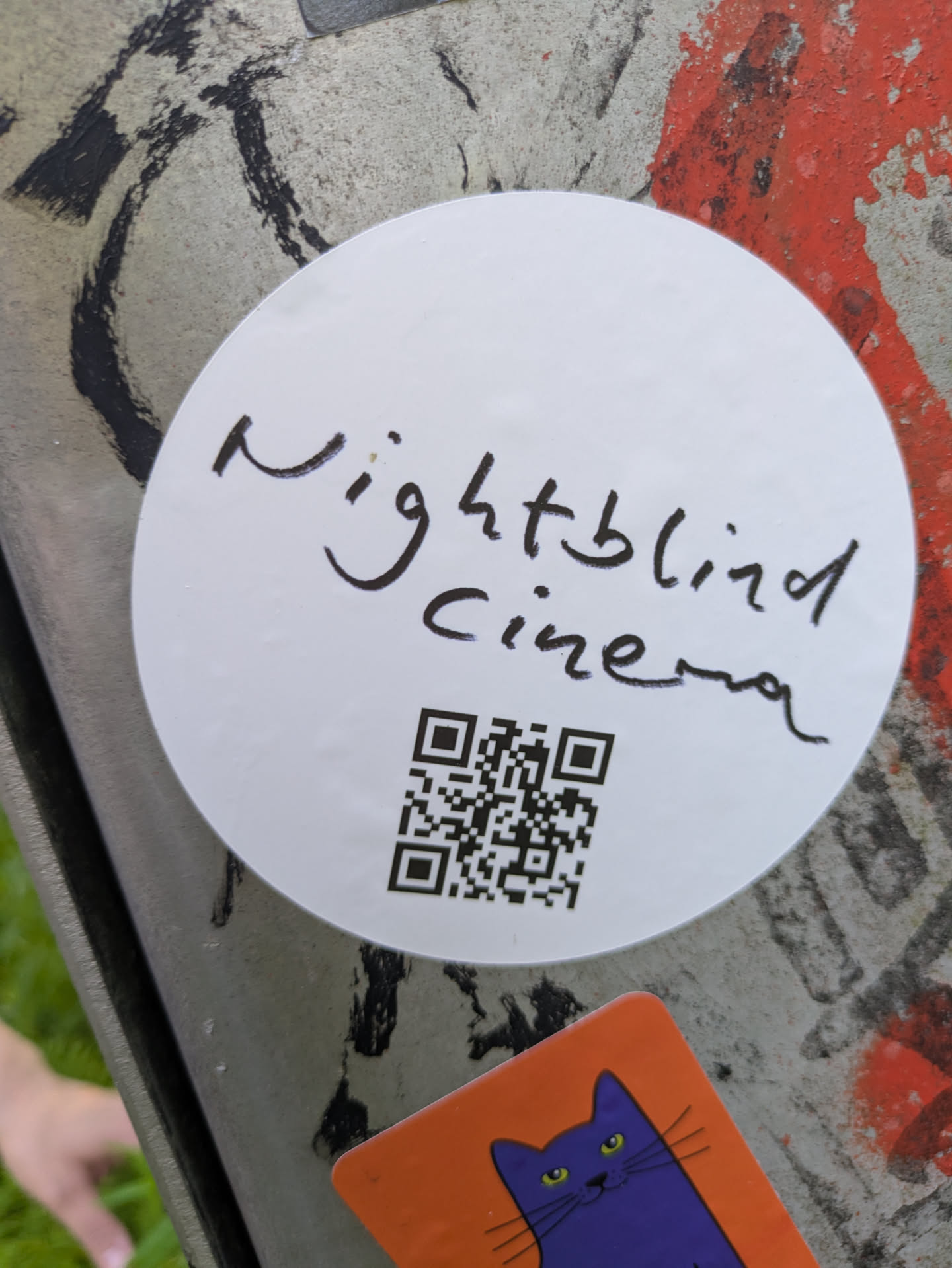 The central element is a white, circular sticker affixed to a weathered, multi-colored surface that appears to be grey metal with patches of peeling red and black paint. The sticker itself is clean and bright white. In the upper half, the words "Nightblind cinema" are written in a distinctive, flowing black script. Below the text, a black QR code is centrally placed, occupying the lower portion of the sticker. The overall impression is minimalist and direct, aiming to direct viewers to the linked content.