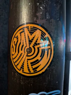 Street sticker The sticker is circular with a black border. The interior is predominantly bright orange with bold black lines forming an abstract, geometric pattern. The design features concentric circles and radiating lines that create a sense of depth and complexity. The overall aesthetic is graphic and eye-catching.