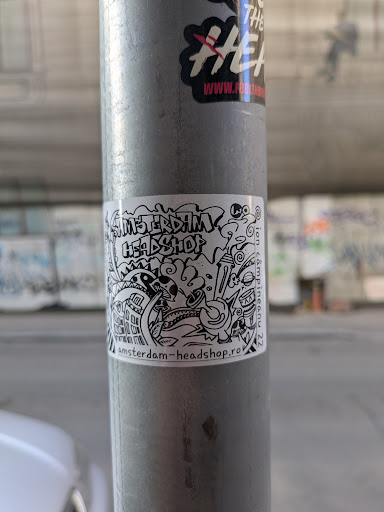 A black and white sticker with a doodle-like drawing and the text "Amsterdam Headshop" and "amsterdam-headshop.ro". The drawing includes elements like a mushroom cloud, various monsters, buildings, and smoking paraphernalia.