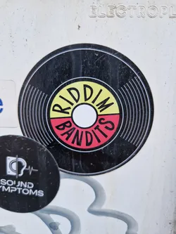 Street sticker A round sticker mimicking a vinyl record. The label features the text "RIDDIM BANDITS" in a stylized font with a color scheme of yellow, red, and white on a black background.  The sticker shows some signs of wear.