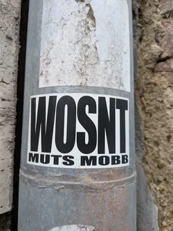 A rectangular white sticker featuring bold black text. The word 'WOSNT' is written in a tall, condensed font above the smaller text 'MUTS MOBB'. It is affixed to a grey metal pipe.