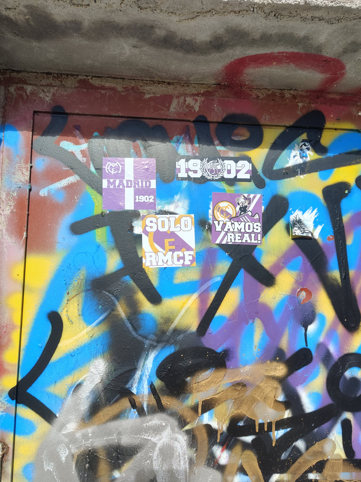 A rectangular sticker with a purple background, featuring a cartoon character in a white shirt and black shorts, wearing a red scarf, in the act of kicking a black and white football. A prominent white and black sneaker with three stripes is visible. In the top left corner, a small Real Madrid crest with a crown and 'M' is present. The lower half of the sticker has white diagonal stripes, and bold white text reads 'VAMOS REAL!' with an exclamation mark.