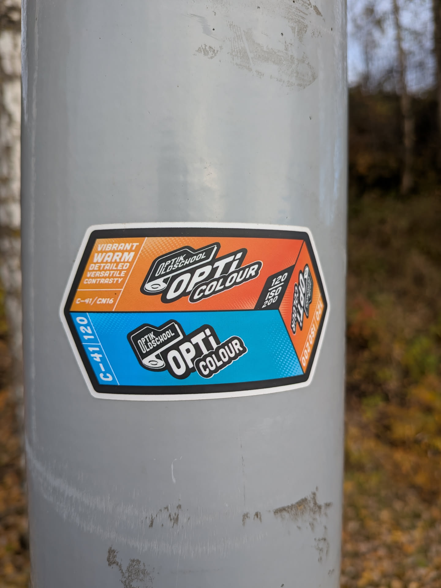 The sticker is hexagonal with rounded corners and a white border. It is divided into two main sections: the top half is orange and the bottom half is blue. The top section features text in white and orange, including 'VIBRANT', 'WARM', 'DETAILED', 'VERSATILE', and 'CONTRASTY'. Below this, in a smaller orange box, is 'C-41/CN16'. The top right of the orange section has a logo with 'OPTIK OLDSCHOOL' above 'OPTI COLOUR' in bold black and white lettering, with a stylized representation of a film reel. The bottom blue section has 'C-41/120' in white text on the left. The bottom right of the blue section has a similar logo with 'OPTIK OLDSCHOOL' above 'OPTI COLOUR'. There are also numerical indicators on the right side of the sticker: '120', 'ISO', '200' in the orange section, and '120 ISO 200' in the blue section, suggesting film speed and format.