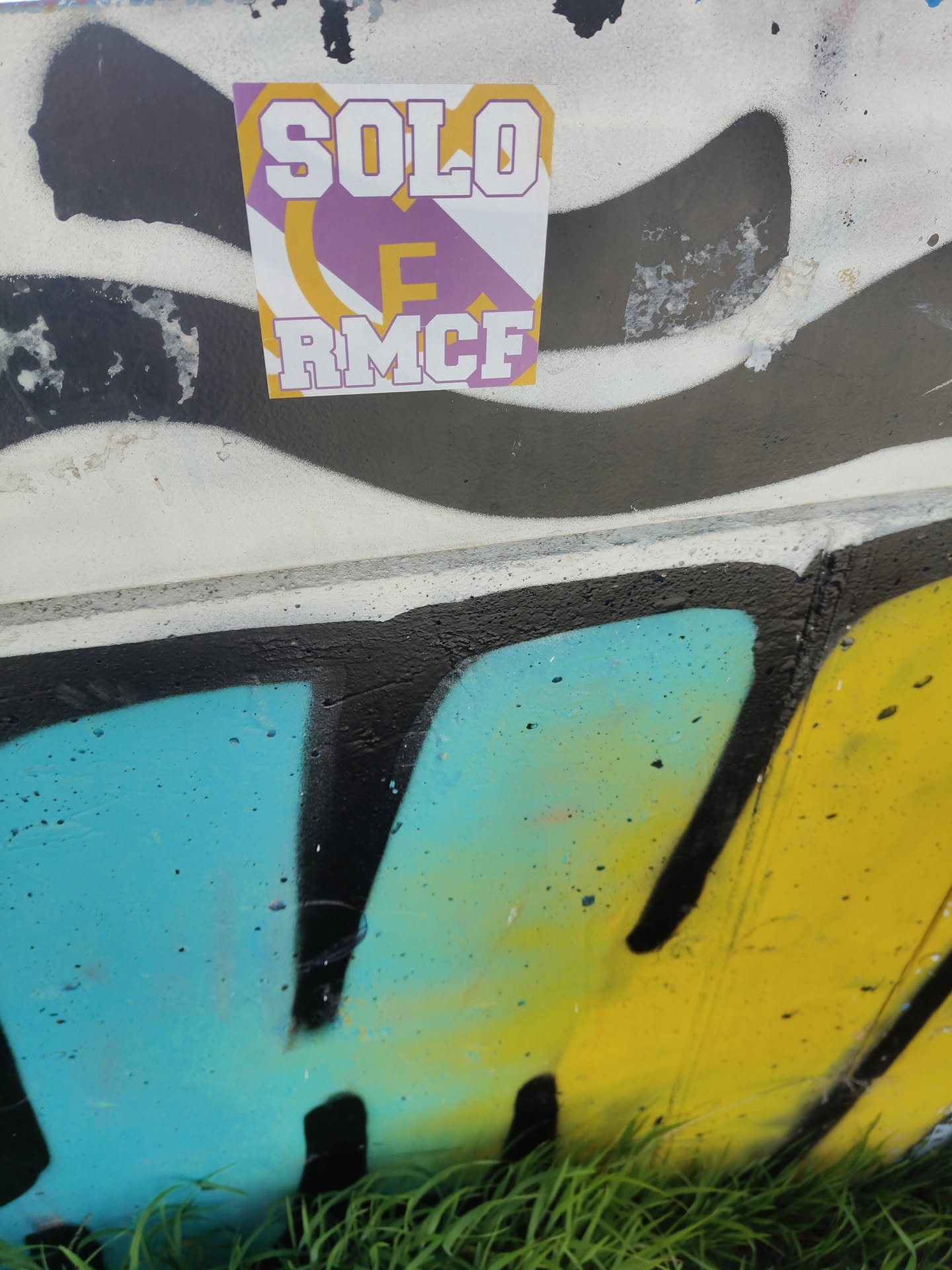 A roughly square-shaped sticker featuring bold, block-style text. The top line reads 'SOLO' in white letters with a purple outline. Below it, a smaller 'E' is rendered in yellow with a purple outline. The bottom line displays 'RMCF' in white letters with a purple outline. The background of the sticker is a dynamic pattern of purple and yellow curved or crescent shapes against a white base. The sticker is affixed to a concrete surface adorned with black, white, blue, and yellow graffiti.