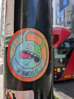 Street sticker A circular sticker featuring a cartoonish, light blue-green creature with a distinctive, stitched-up appearance. The creature has two small, light green horns or ears. Its left eye is a white 'X' stitch, while its right eye is a small yellow circle with a black pupil. A wide-open mouth reveals a bright pink tongue, white teeth, and a dark blue interior, suggesting a scream or laugh. Dashed lines resembling stitches are visible across its head, particularly on the right side of its face and below its left eye. The creature is set against a background that is split between solid orange-red on the right and light yellow with dark blue diagonal stripes on the left. The sticker has an outer orange-red border, followed by thin green and blue inner borders. The word 'IRONMOULD' is written in dark blue capital letters along the bottom curve of the inner circle.