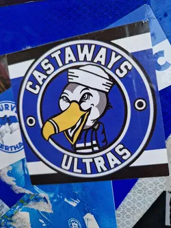 Street sticker A blue and white circular sticker with the words "Castaways Ultras" and an illustration of an albatross in a sailor's outfit. The albatross is wearing a white sailor's hat and a blue and white striped scarf. The sticker has a clean and simple design.
