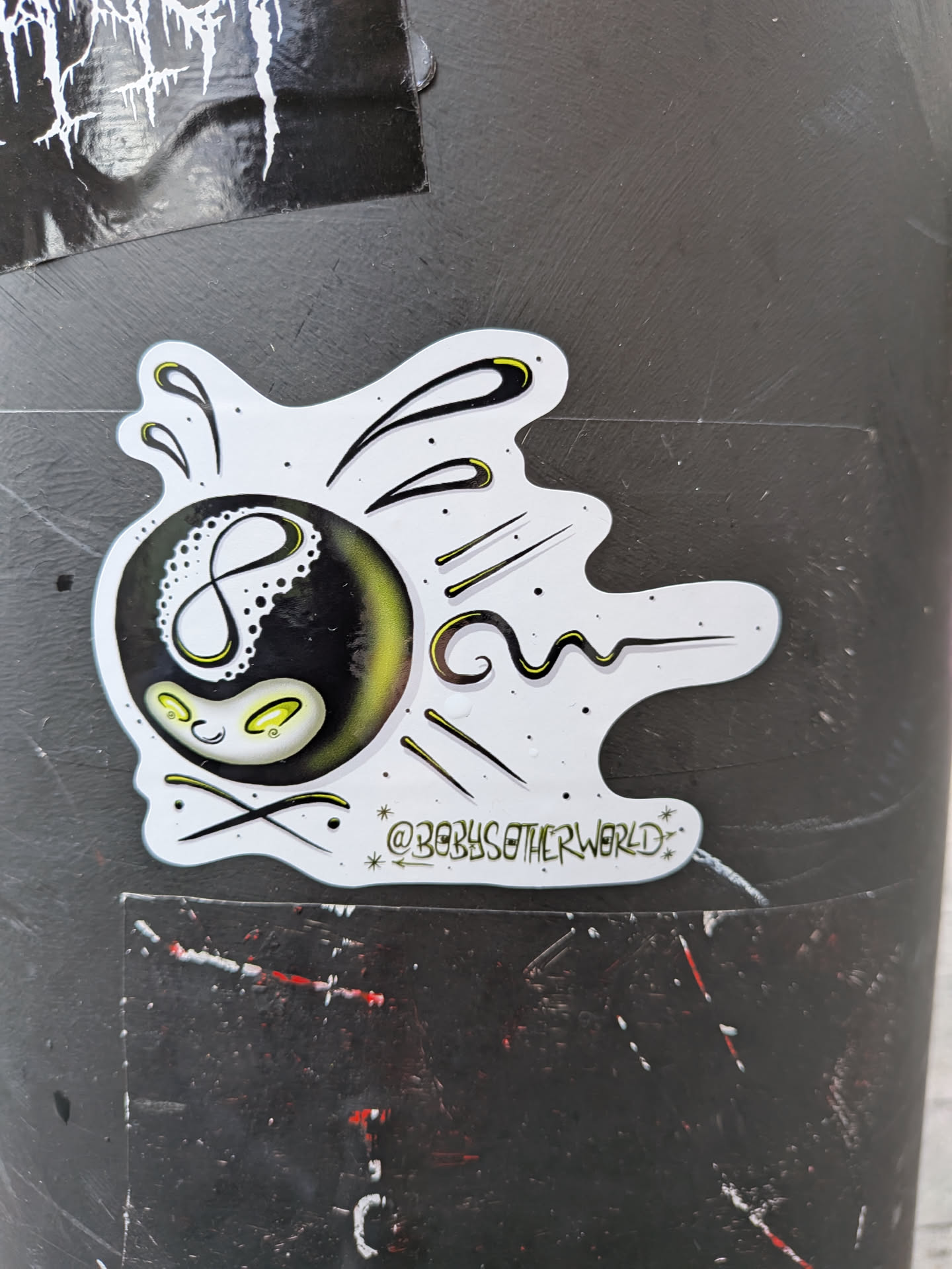 An irregularly shaped sticker with a white outline, featuring a stylized black 8-ball as its central element. The '8' on the ball is white and has a delicate, dotted, lace-like pattern on its left side. Below the '8', a small, anthropomorphic face is depicted with two glowing yellow-green oval eyes and a subtle, curved smile. The bottom portion of the 8-ball also emits a soft yellow-green glow. Emanating from the 8-ball are various black lines and dots, some accented with yellow-green, suggesting motion or energy against the white background. Small black speckles are scattered across the white areas of the sticker. Below the main design, the text '@BOBYSOTHERWORLD' is written in a stylized font, flanked by small starbursts.