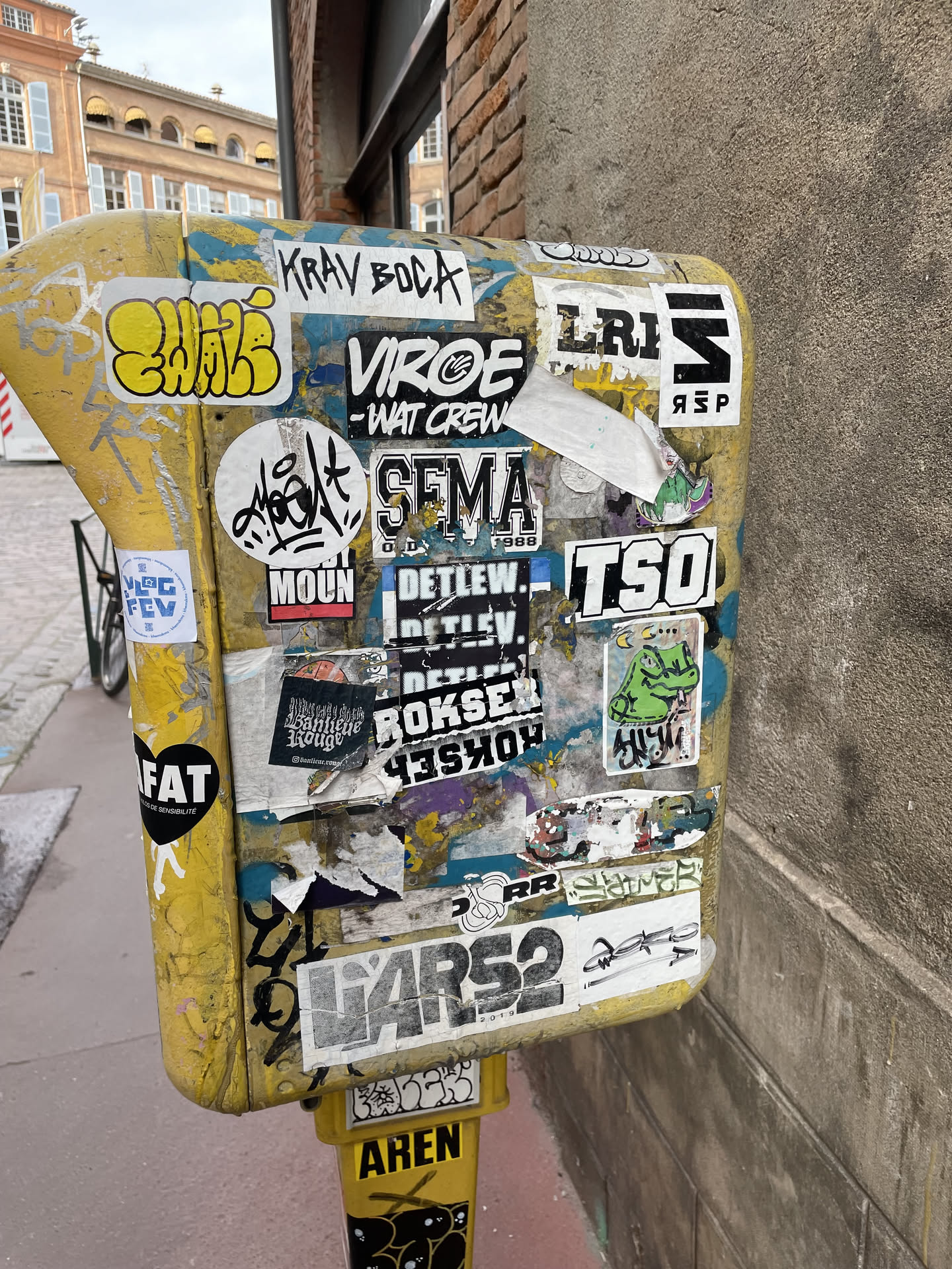 Street sticker The sticker is located at the center of a yellow, cylindrical object, likely a post or bollard, which is heavily covered in various stickers and graffiti. The stickers are layered and overlapping, creating a dense visual texture. Prominent stickers include bold, stylized lettering in black and white, such as 'KRAV BOCA', 'VIROE -WAT CREW', 'SEMA 1988', 'TSO', and 'FAT'. There are also more abstract graphic elements, including a yellow bubble-letter tag, a white circular sticker with black graffiti-style writing, and a small green cartoon frog-like creature with a crescent moon. Some stickers are torn or peeling, revealing layers underneath or the yellow surface of the post. The overall impression is one of a dynamic and evolving urban canvas.