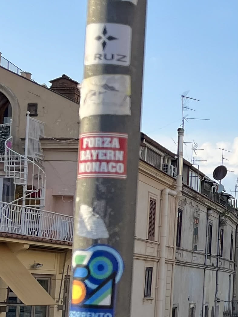 The sticker is rectangular with a bright red background and bold white capitalized text. The text is arranged in three lines: 'FORZA' on the top, 'BAYERN' in the middle, and 'MONACO' at the bottom. The sticker appears slightly worn around the edges, indicating it's a physical adhesive item on a grey pole.