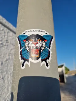 A sticker featuring a traditional-style tattoo of a panther head within butterfly wings. The panther is black with yellow eyes and red mouth details, the wings are teal. The artist's Instagram handle is on the sticker: @curledvurptattoo