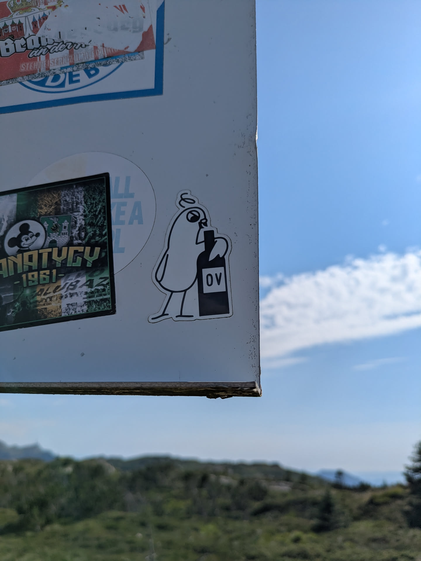 A black and white cartoon sticker depicting a stylized, blob-like character with an expressive face and what appears to be a halo or curly hair on its head. The character is holding a dark bottle with the letters 'OV' clearly visible on its label. The character has small, simple legs and feet, and round eyes with a pointed nose or beak. It's stuck on a light-colored pole or sign, with a clear blue sky and distant green landscape in the background.