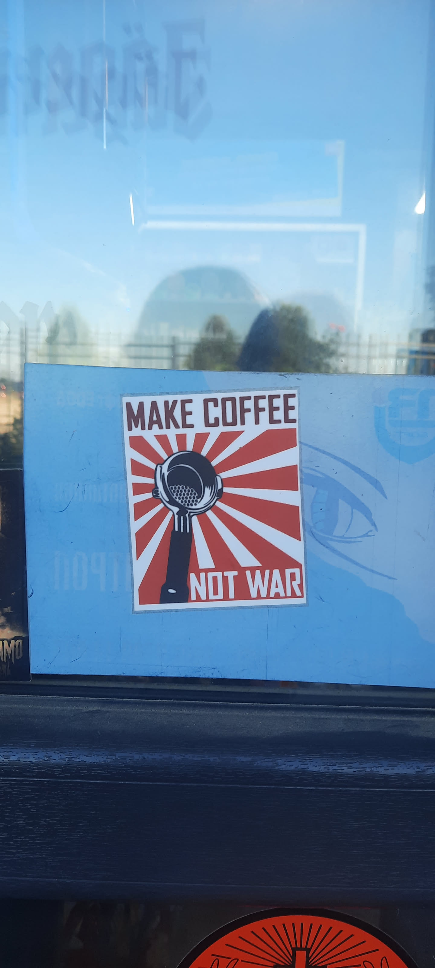 Street sticker A rectangular sticker featuring a central black and white illustration of an espresso machine portafilter with a coffee basket, set against a vibrant background of red rays radiating from the center on a white base, reminiscent of the Japanese Rising Sun flag. Above the portafilter, the text 'MAKE COFFEE' is displayed in dark red/brown letters on a white rectangular banner. Below the portafilter, the text 'NOT WAR' is similarly displayed. The entire design has a thin white border.