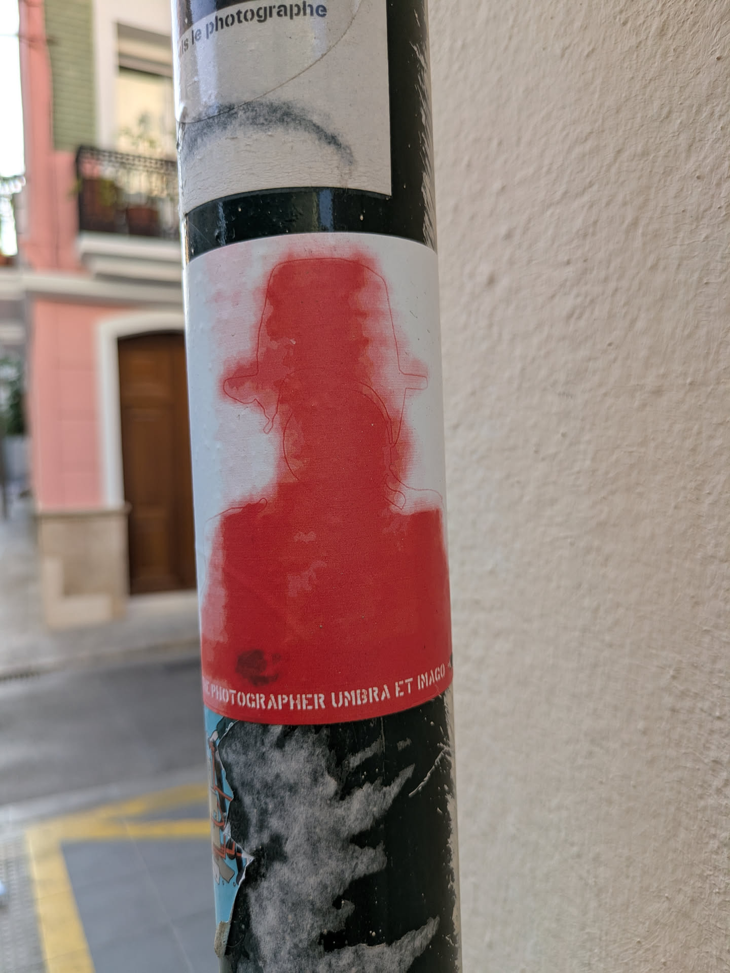 A red silhouette sticker featuring a figure with a hat, adhered to a pole. The sticker has the text 'PHOTOGRAPHER UMBRA ET IMAGO' on it.