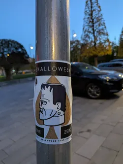 Street sticker The sticker features a profile illustration of Frankenstein's monster's head. The monster has dark hair, a prominent forehead, and stitched details on its neck. The illustration is rendered in black and white with a golden-brown background behind the head, contained within a triangular shape. Below the monster's head, there is a black rectangular bar with white text and a date. The sticker has a white border and appears to be adhered to a metal pole.
