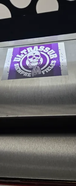 Street sticker A rectangular sticker featuring a prominent white skull graphic centered on a vibrant purple background. The skull is depicted wearing a scarf or bandana around its neck, with the letters 'US' or 'U8' faintly visible on it. Two concentric white rings encircle the skull, and around the outer ring, the white text 'ULTRASSUR SEMPRE FIELES' is arranged. The purple background has a distressed, almost splattered white effect along its left and right edges, giving the sticker a rugged and worn appearance. The overall design is graphic and uses a strong two-tone purple and white color scheme.