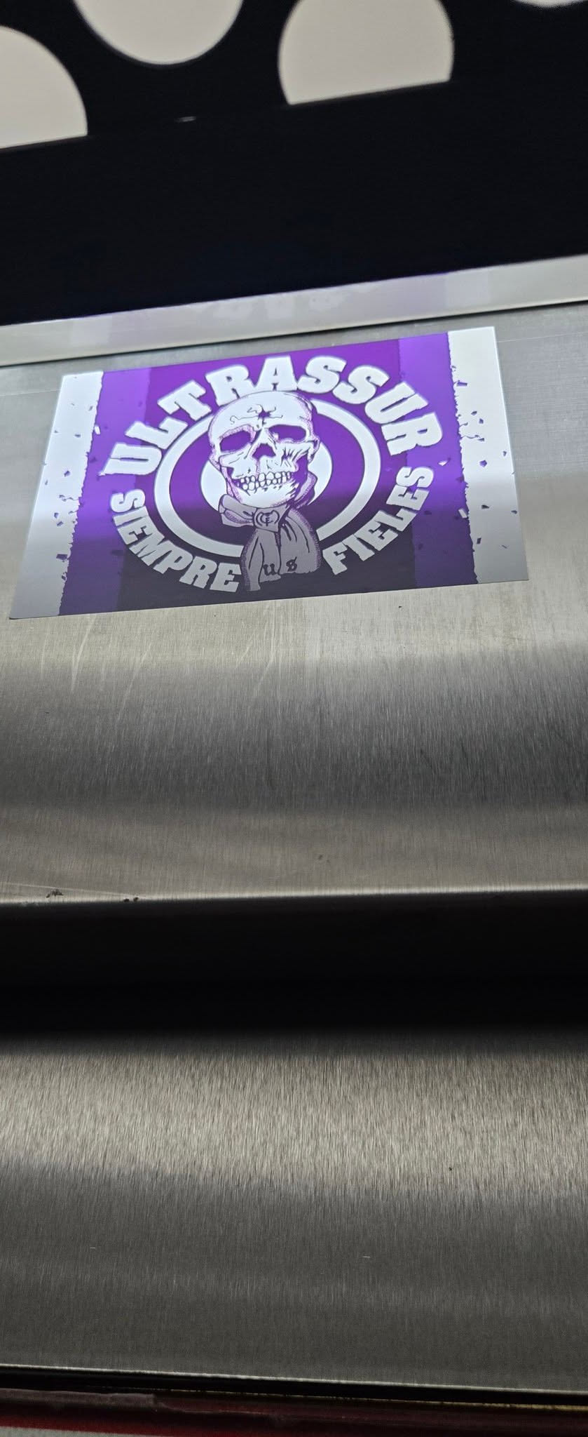 A rectangular sticker featuring a prominent white skull graphic centered on a vibrant purple background. The skull is depicted wearing a scarf or bandana around its neck, with the letters 'US' or 'U8' faintly visible on it. Two concentric white rings encircle the skull, and around the outer ring, the white text 'ULTRASSUR SEMPRE FIELES' is arranged. The purple background has a distressed, almost splattered white effect along its left and right edges, giving the sticker a rugged and worn appearance. The overall design is graphic and uses a strong two-tone purple and white color scheme.