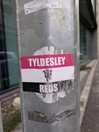 A sticker found on a metal pole. The sticker is a tricolor with the words 'TYLDESLEY' on a pink background at the top, a white middle section with a stylized devil logo and 'REDS' in white letters on a black background at the bottom.