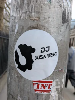 Street sticker A circular, black and white sticker featuring a minimalist design. The left side displays a black silhouette of a person's head in profile, facing left, wearing large headphones. The headphones are also rendered in black, creating a negative space cutout within the head's outline. To the right of the silhouette, the text 'DJ JUSA BEAT' is printed in a clean, sans-serif black font, with 'DJ' stacked above 'JUSA BEAT'. The overall aesthetic is clean and focused on music branding.