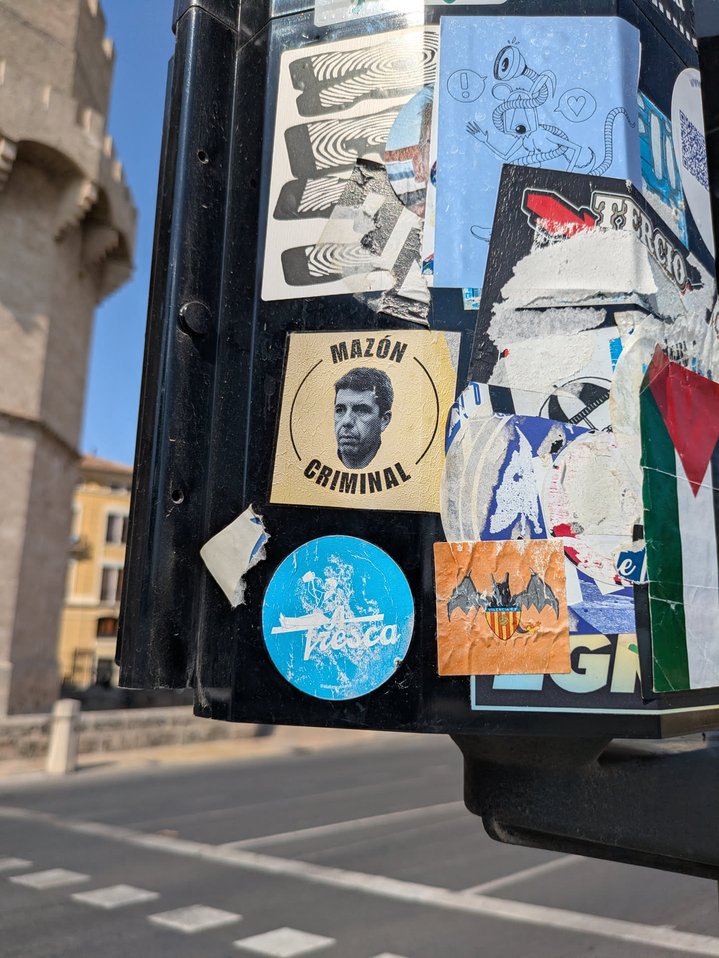 A close-up of a black pole covered with various stickers. The most prominent stickers include a circular sticker with a portrait and the text "MAZON CRIMINAL", and a round blue sticker with the word "Fresco" in white script. Other stickers include abstract designs and symbols, creating a dense and layered effect.
