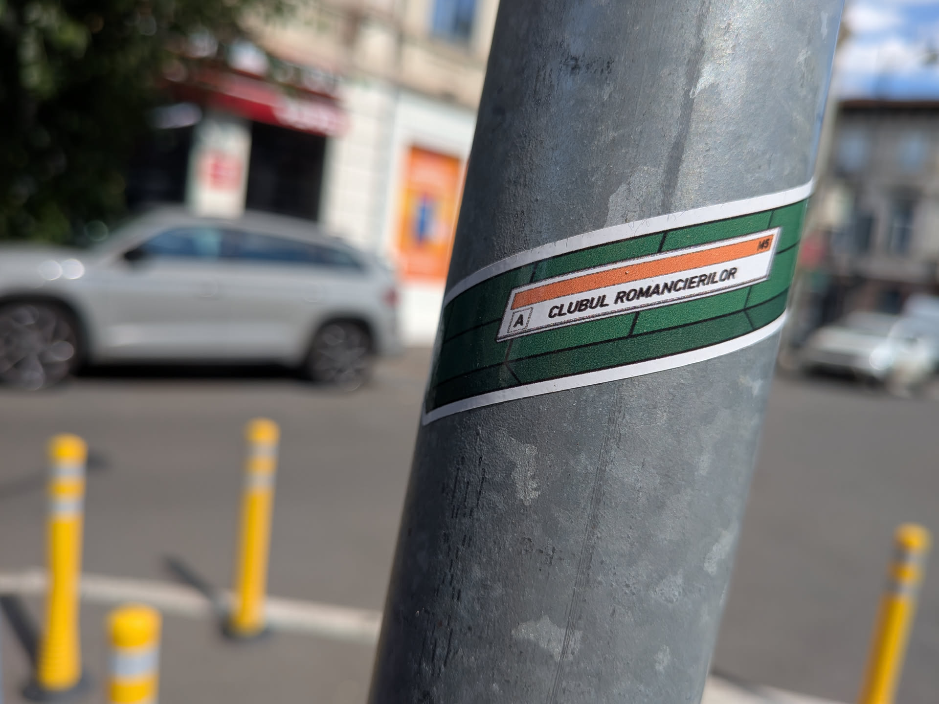 A rectangular sticker mimicking a Bucharest metro sign. It features a dark green background with horizontal lines and a central white label containing an orange stripe, the letter 'A', the text 'CLUBUL ROMANCIERILOR', and the number '145'.
