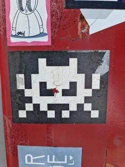 A retro-style pixel art Space Invader sticker is affixed to a worn, red surface. The sticker shows some signs of wear and tear, adding to its vintage aesthetic. The pixelated design is classic, reminiscent of early video games.