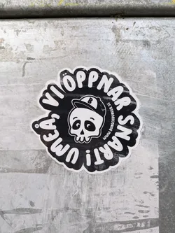 Street sticker Black and white round sticker with a cartoon skull wearing a baseball cap in the center.  The text around the skull says  "VI ÖPPNAR SNART I UMEA", which translates from Swedish to "WE OPEN SOON IN UMEA". A small website address is visible but unreadable. 