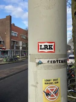 Street sticker The central sticker is white with a red, scalloped border. Inside the border, the letters 'LRK' are prominently displayed in bold, black, sans-serif font. Directly below this sticker, another sticker is visible, featuring a bright yellow background. This sticker has text at the top reading 'LOBBY ERUIT BURGERS ERIN' in black and orange. Below the text is the symbol for Extinction Rebellion, depicted as a circle with a triangle inside, stylized with an hourglass shape. Two birds, one appearing as a skeleton and the other a live bird, are pecking at each other within the symbol.