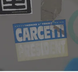 Street sticker The sticker is rectangular with a blue background at the top and a pale yellow background at the bottom. The top section features white text that reads 'THOMAS J. "TOMMY"' with a row of white stars above it. Below this, in large, bold white letters, is the name 'CARCETTI'. The bottom section has the word 'PRESIDENT' in large, faded white letters. The sticker shows signs of wear and tear, with cracks and peeling in the material.