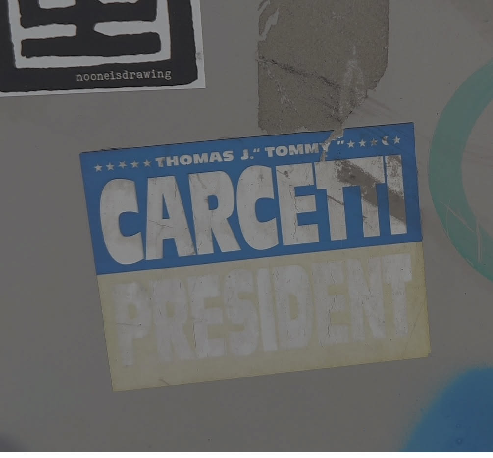 The sticker is rectangular with a blue background at the top and a pale yellow background at the bottom. The top section features white text that reads 'THOMAS J. "TOMMY"' with a row of white stars above it. Below this, in large, bold white letters, is the name 'CARCETTI'. The bottom section has the word 'PRESIDENT' in large, faded white letters. The sticker shows signs of wear and tear, with cracks and peeling in the material.