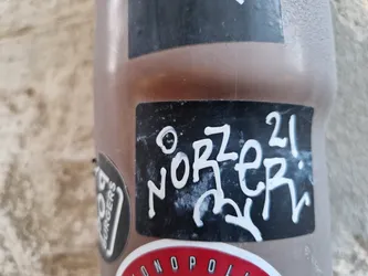 Street sticker A black sticker with white graffiti-style text reading "Oz Norer 21". The sticker is attached to a brown object, possibly a bottle or container.