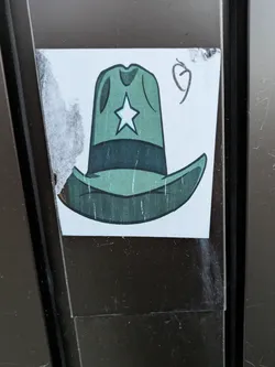 Street sticker A sticker featuring a cartoon illustration of a green cowboy hat with a white star on the crown. The sticker appears to be hand-drawn or digitally created with a simple, slightly rustic style. It's affixed to a dark brown surface, showing some signs of wear and tear around the edges.