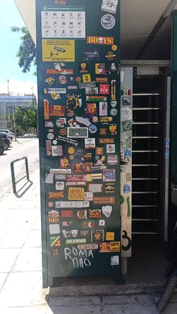 The image centers on a dark green utility box or cabinet, completely covered in a dense, overlapping collection of stickers and some graffiti. The stickers vary greatly in size, shape, and design, predominantly featuring football club emblems, fan group logos, and slogans related to AS Roma and SS Lazio, with some references to Olympiakos and Panathinaikos. Common motifs include wolves, eagles, skulls, and various crests. Colors are diverse, with prominent reds, yellows, whites, and blacks. The overall appearance is chaotic yet expressive, reflecting passionate fan culture. Below the stickers, white graffiti spells out 'ROMA PAO'. At the top, there are official signs in Greek and English about prohibited items and CCTV surveillance.
