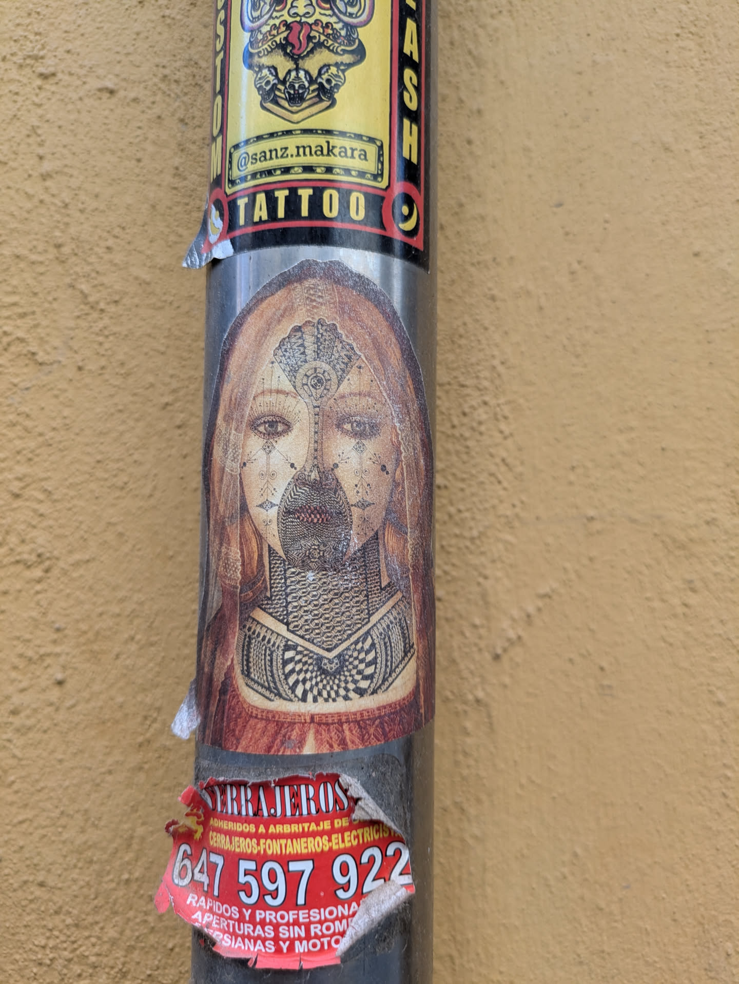 The sticker depicts a woman's face with tattoos, placed on a pole alongside other stickers. Above it, there's a sticker of a logo with text that reads "@sanz.makara TATTOO". Below the sticker is another one that says "cerrajeros"
