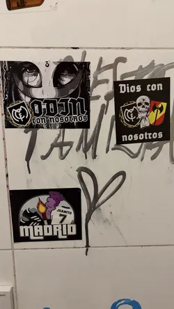 Street sticker This sticker features a striking grayscale illustration of a character's face, dominated by large, stylized anime-like eyes with prominent eyelashes and dark pupils. The character has dark, possibly braided or dreadlocked hair framing the face. Below the face, bold, blocky text reads 'ODIN' in white, with smaller, more gothic-style text beneath it saying 'con nosotros'. To the left of the 'ODIN' text is a stylized crest, predominantly black and white with gold accents, resembling a football club emblem (specifically, it strongly evokes the Real Madrid C.F. crest). The overall aesthetic is dark, mysterious, and urban, with a strong graphic novel or street art feel.