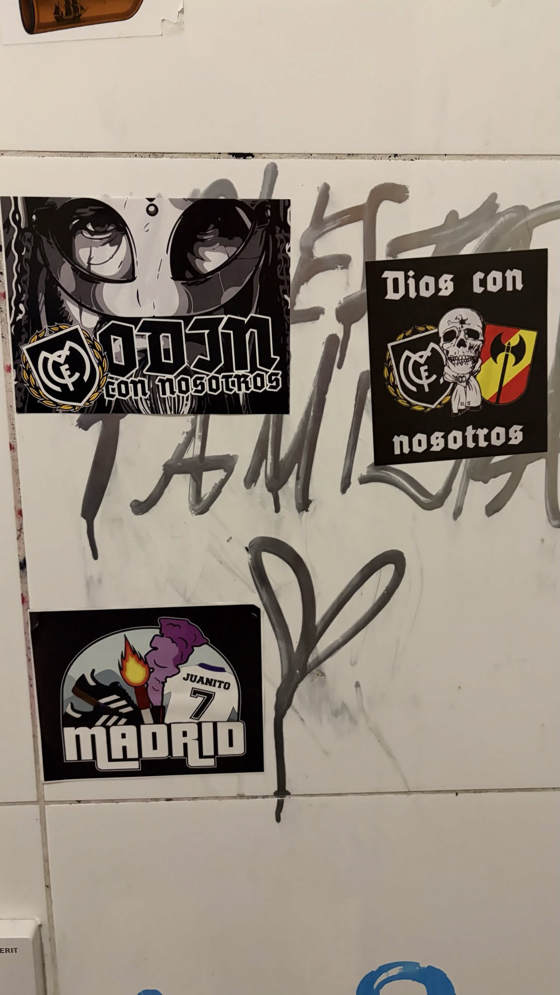 This sticker features a striking grayscale illustration of a character's face, dominated by large, stylized anime-like eyes with prominent eyelashes and dark pupils. The character has dark, possibly braided or dreadlocked hair framing the face. Below the face, bold, blocky text reads 'ODIN' in white, with smaller, more gothic-style text beneath it saying 'con nosotros'. To the left of the 'ODIN' text is a stylized crest, predominantly black and white with gold accents, resembling a football club emblem (specifically, it strongly evokes the Real Madrid C.F. crest). The overall aesthetic is dark, mysterious, and urban, with a strong graphic novel or street art feel.